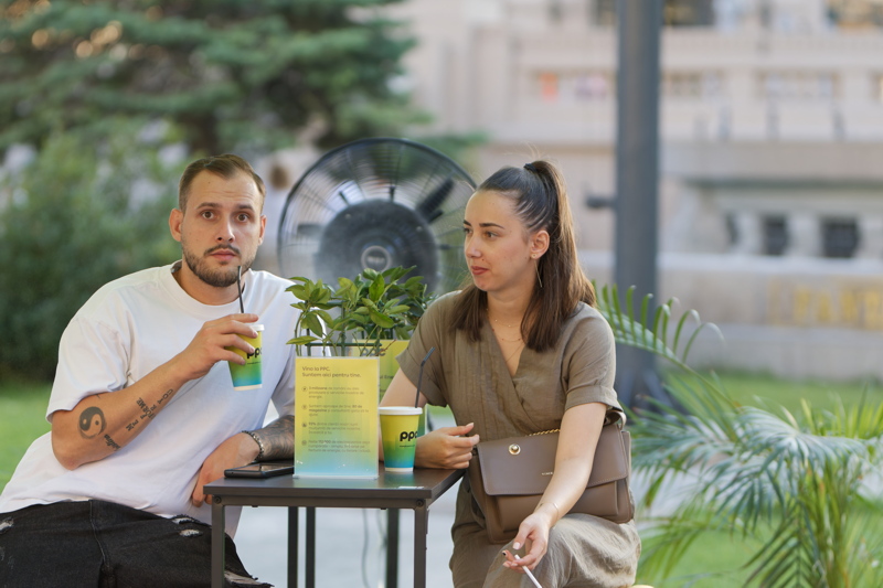 Two people sitting at a small black metal table on an outdoor terrace, with the woman on the right holding a brown purse while conversing